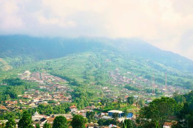 Kabarık beyaz bulutlarla çevrili yemyeşil bir Merbabu dağının nefes kesici manzarası ve açık mavi gökyüzünün arka planına kurulmuş tuhaf bir köy.