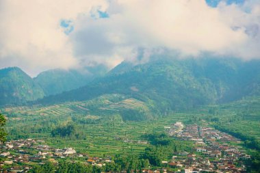 Kabarık beyaz bulutlarla çevrili yemyeşil bir Merbabu dağının nefes kesici manzarası ve açık mavi gökyüzünün arka planına kurulmuş tuhaf bir köy.