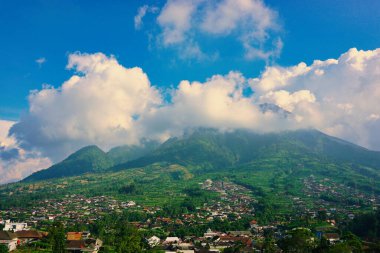 Kabarık beyaz bulutlarla çevrili yemyeşil bir Merbabu dağının nefes kesici manzarası ve açık mavi gökyüzünün arka planına kurulmuş tuhaf bir köy.