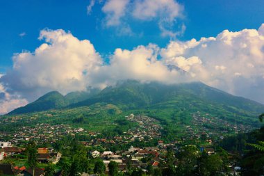 Kabarık beyaz bulutlarla çevrili yemyeşil bir Merbabu dağının nefes kesici manzarası ve açık mavi gökyüzünün arka planına kurulmuş tuhaf bir köy.