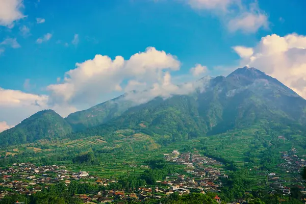Kabarık beyaz bulutlarla çevrili yemyeşil bir Merbabu dağının nefes kesici manzarası ve açık mavi gökyüzünün arka planına kurulmuş tuhaf bir köy.