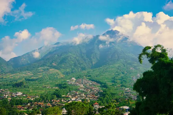 Kabarık beyaz bulutlarla çevrili yemyeşil bir Merbabu dağının nefes kesici manzarası ve açık mavi gökyüzünün arka planına kurulmuş tuhaf bir köy.