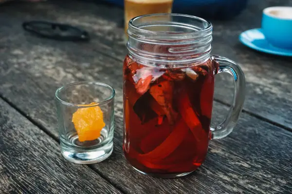 Bir kavanoz java bitkisel çayı ya da Wedang Uwuh 'a içinde bal olan bir bardak eşlik eder, ahşap bir yüzeye kurulmuş, samimi ve doğal bir içecek sahnesini canlandırır.