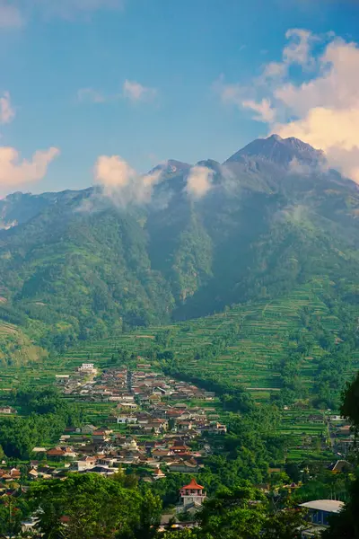 Kabarık beyaz bulutlarla çevrili yemyeşil bir Merbabu dağının nefes kesici manzarası ve açık mavi gökyüzünün arka planına kurulmuş tuhaf bir köy.