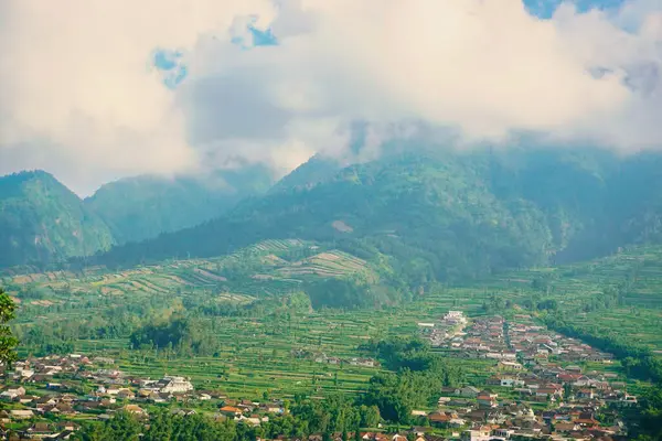 Kabarık beyaz bulutlarla çevrili yemyeşil bir Merbabu dağının nefes kesici manzarası ve açık mavi gökyüzünün arka planına kurulmuş tuhaf bir köy.