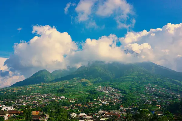 Kabarık beyaz bulutlarla çevrili yemyeşil bir Merbabu dağının nefes kesici manzarası ve açık mavi gökyüzünün arka planına kurulmuş tuhaf bir köy.