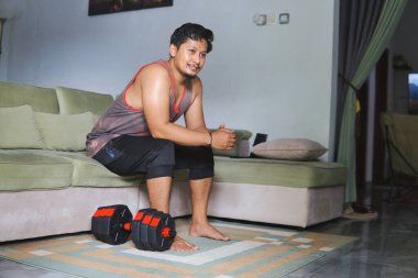 Oturma odasında halterle egzersiz yapan bir adam kanepeye oturur ve düşünür.