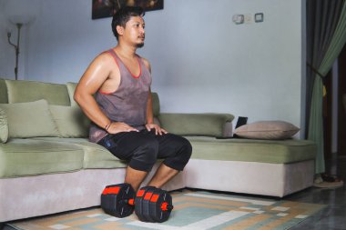 Oturma odasında halterle egzersiz yapan bir adam kanepeye oturur ve düşünür.