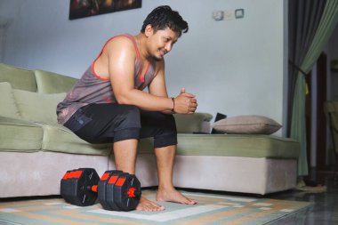 Oturma odasında halterle egzersiz yapan bir adam kanepeye oturur ve düşünür.