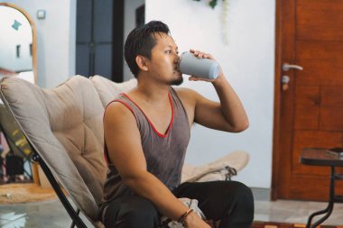 Bir erkek egzersiz seansından sonra oturma odasında dinlenir. Sütlü proteinini yudumlayarak evde sağlıklı ve aktif bir yaşam tarzını temsil eder.