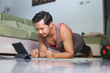 Yoga minderi üzerinde tahta egzersizi yapan bir adam, bir tablette sergilenen online bir egzersiz yaparken, rahat bir ev ortamında spor yapmaya kendini adamış.