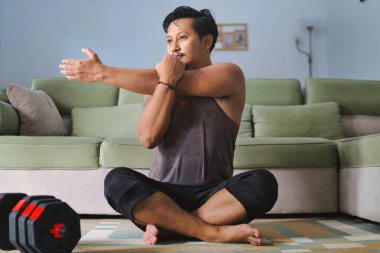 Oturma odasının zemininde oturan bir adam, rahat ve fitness odaklı ev ortamını sergiliyor, ayarlanabilir halterler yakınlarda görülebiliyor, ilham verici bir ev uygunluğu atmosferi yaratıyor..