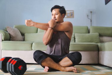 Oturma odasının zemininde oturan bir adam, rahat ve fitness odaklı ev ortamını sergiliyor, ayarlanabilir halterler yakınlarda görülebiliyor, ilham verici bir ev uygunluğu atmosferi yaratıyor..