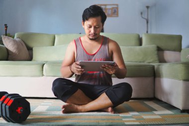 Samimi bir oturma odasında halının üzerinde oturan bir adam, spor seansına hazırlanırken, spor malzemeleriyle çevrili, tablet kullanıyor. Kurulum sağlık ve kapalı alandaki faaliyetleri iletir.