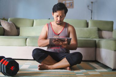 Bir adam halı kaplı bir zeminde bağdaş kurarak oturuyor, bir tablet cihazına odaklanmış, yanında ayarlanabilir bir halterle, fitness, verimlilik ve modern teknolojiyi vurguluyor..