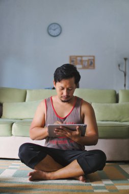 Bir adam halı kaplı bir zeminde bağdaş kurarak oturuyor, bir tablet cihazına odaklanmış, yanında ayarlanabilir bir halterle, fitness, verimlilik ve modern teknolojiyi vurguluyor..