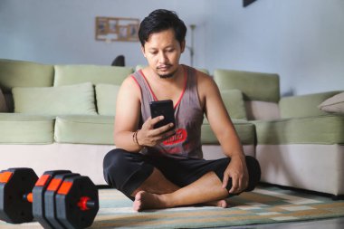 Günlük giysiler içinde bir adam oturma odasında yerde oturuyor, akıllı telefonunu kullanıyor. Dambıllar, modern bir ev ortamında fitness ya da rahatlama teması izlenimi veren yakınlara yerleştirilmiştir..