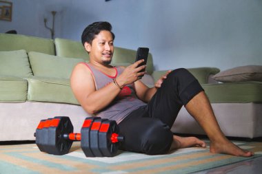 Spor kıyafeti giymiş bir adam oturma odasında yerde oturuyor, akıllı telefonunu tutuyor, yanında bir halterle antreman yaptıktan sonra dinleniyor. Rahat ve rahat bir egzersiz sonrası anı yakalıyor.