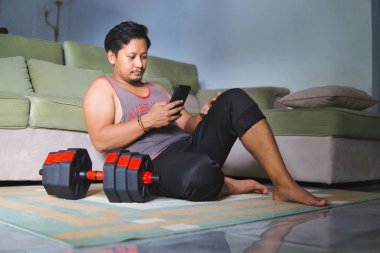 Spor kıyafeti giymiş bir adam oturma odasında yerde oturuyor, akıllı telefonunu tutuyor, yanında bir halterle antreman yaptıktan sonra dinleniyor. Rahat ve rahat bir egzersiz sonrası anı yakalıyor.