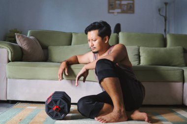 Egzersizden sonra dinlenen bir adam evinin oturma odasında dambılları kaldırıyor.