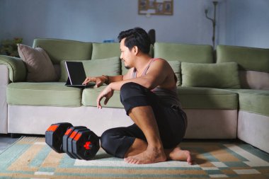 Bir adam rahat bir ev ortamında yerde oturuyor, dijital tablet kullanıyor ve ayarlanabilir dambıllarla spor yapıyor.