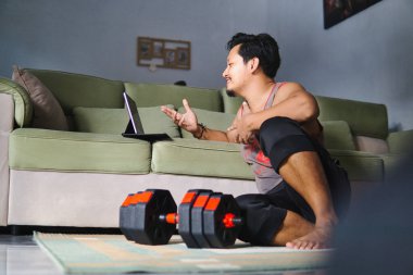 Bir adam oturma odasında yerde oturuyor, tablet kullanarak sanal bir seansa katılıyor. Yakınlarda, bir çift dambıl egzersiz ya da egzersiz bağlamını belirtir, spor ve teknolojiyi vurgular..