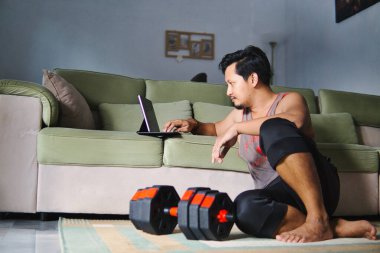 Bir adam rahat bir ev ortamında yerde oturuyor, dijital tablet kullanıyor ve ayarlanabilir dambıllarla spor yapıyor.