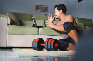 Bir adam oturma odasında yerde oturuyor, tablet kullanarak sanal bir seansa katılıyor. Yakınlarda, bir çift dambıl egzersiz ya da egzersiz bağlamını belirtir, spor ve teknolojiyi vurgular..