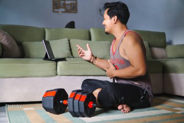 Sıradan bir ortamda, sanal bir fitness aktivitesine katılan bir adam. Ağırlıkların yanında oturmak ve elektronik bir cihazla meşgul olmak, sağlık, ev konforu ve dijital bağlantıyı vurgulamak