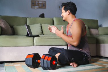 Sıradan bir ortamda, sanal bir fitness aktivitesine katılan bir adam. Ağırlıkların yanında oturmak ve elektronik bir cihazla meşgul olmak, sağlık, ev konforu ve dijital bağlantıyı vurgulamak