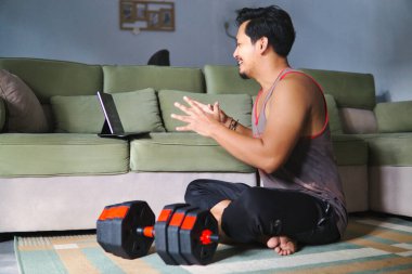 Sıradan bir ortamda, sanal bir fitness aktivitesine katılan bir adam. Ağırlıkların yanında oturmak ve elektronik bir cihazla meşgul olmak, sağlık, ev konforu ve dijital bağlantıyı vurgulamak
