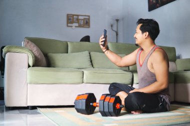 Akıllı telefonuyla yerde oturan bir adam, evde yeşil bir kanepenin yanında egzersiz yapıyor. Sahne, kişisel yaşam alanında teknoloji ve konfor içeren modern fitness rutinlerini vurguluyor.