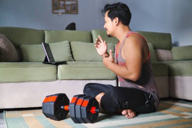 Sıradan bir ortamda, sanal bir fitness aktivitesine katılan bir adam. Ağırlıkların yanında oturmak ve elektronik bir cihazla meşgul olmak, sağlık, ev konforu ve dijital bağlantıyı vurgulamak