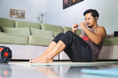 Sıcak bir oturma odasında oturma egzersizi yapan bir adam, sağlıklı bir yaşam tarzı için evde çalışma alışkanlıkları sergiliyor. Spor kıyafetleri giyiyor ve kulaklıkla müzik dinlerken paspas kullanıyor.