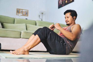 Sıcak bir oturma odasında oturma egzersizi yapan bir adam, sağlıklı bir yaşam tarzı için evde çalışma alışkanlıkları sergiliyor. Spor kıyafetleri giyiyor ve kulaklıkla müzik dinlerken paspas kullanıyor.