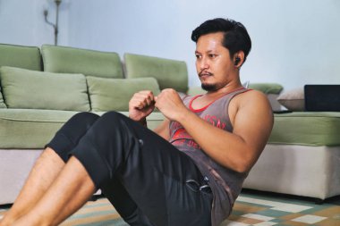 Sıcak bir oturma odasında oturma egzersizi yapan bir adam, sağlıklı bir yaşam tarzı için evde çalışma alışkanlıkları sergiliyor. Spor kıyafetleri giyiyor ve kulaklıkla müzik dinlerken paspas kullanıyor.