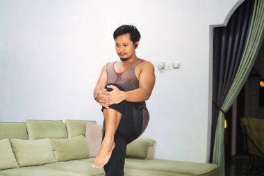 Bir adam evde bir denge ve esneme egzersizi yapar, diğerini göğsüne doğru tutarken bir bacağı üzerinde durur. Sağlığı, fiziksel kondisyonu ve sağlığı teşvik eder.