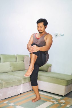 Bir adam evde bir denge ve esneme egzersizi yapar, diğerini göğsüne doğru tutarken bir bacağı üzerinde durur. Sağlığı, fiziksel kondisyonu ve sağlığı teşvik eder.
