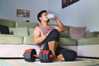 Bir adam egzersiz sonrası oturma odasında dinleniyor, sütlü proteinini yudumluyor ve halterin yanında oturuyor, evde sağlıklı ve aktif bir yaşam tarzını temsil ediyor.
