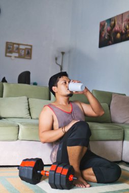 Bir adam egzersiz sonrası oturma odasında dinleniyor, sütlü proteinini yudumluyor ve halterin yanında oturuyor, evde sağlıklı ve aktif bir yaşam tarzını temsil ediyor.