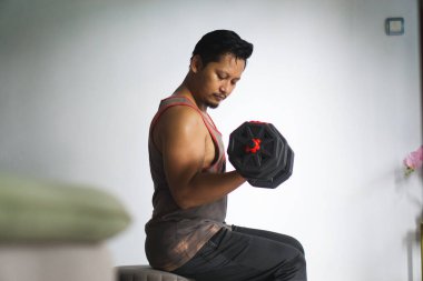 Bir adam evde oturmuş, halterleri kaldırıyor ve günlük ev ortamında güç antrenmanına odaklanıyor.