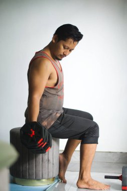 Bir adam evde oturmuş, halterleri kaldırıyor ve günlük ev ortamında güç antrenmanına odaklanıyor.