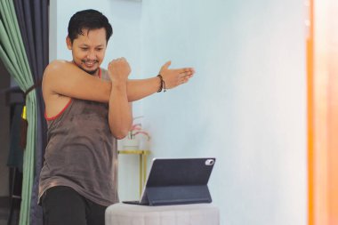 Sıradan bir evde kol esnetme alıştırması yapan bir adam, bir tablette gösterilen çevrimiçi bir çalışmayla etkileşime girer. Odanın düzeni odak ve aktivite duygusunu arttırır.