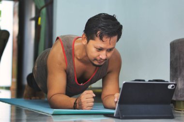 Yoga minderi üzerinde tahta egzersizi yapan bir adam, bir tablette sergilenen online bir egzersiz yaparken, rahat bir ev ortamında spor yapmaya kendini adamış.