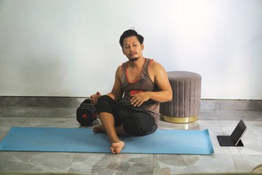 Ev ortamında yoga minderinde oturan bir adam, yanında bir dambıl ve tablet ile birlikte, konforlu ev ortamından spor yapmayı ya da spor yapmayı öneriyor..