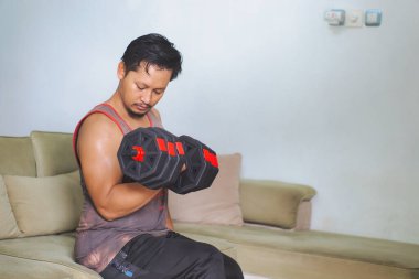 Bir erkek evdeki bir kanepede halterlerle egzersiz yapar, ev ortamında spor ve refahı teşvik eder..