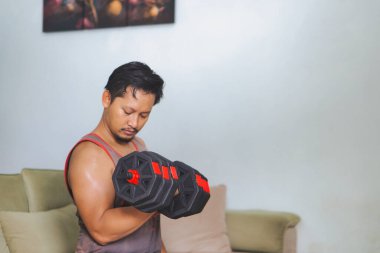 Bir adam evde oturmuş, halterleri kaldırıyor ve günlük ev ortamında güç antrenmanına odaklanıyor.