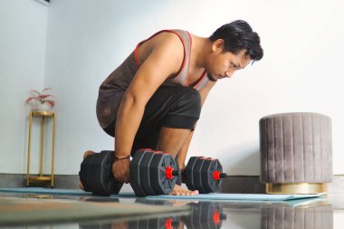 Kol halterleri kullanarak içeride egzersiz yapan motive olmuş bir birey. Bir ev ortamında kendini adamanın, sağlığın ve kendini geliştirmenin özünü yakalar.