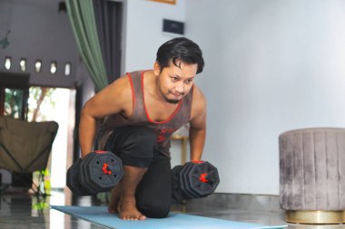 Kol halterleri kullanarak içeride egzersiz yapan motive olmuş bir birey. Bir ev ortamında kendini adamanın, sağlığın ve kendini geliştirmenin özünü yakalar.