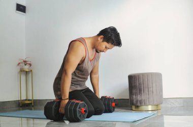 Evde dambıllarla egzersiz yaparken mola veren bir adam.